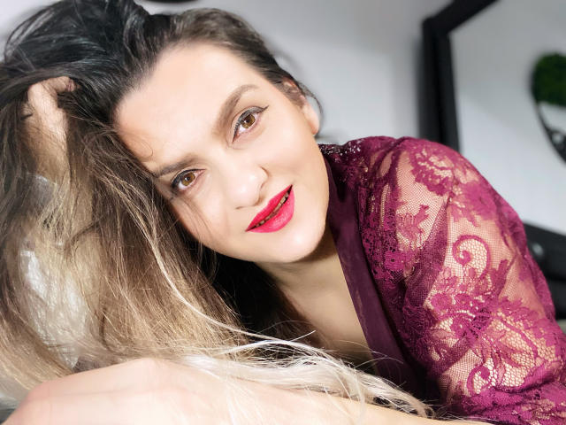 ReveMignoneX com cabelo loiro, batom vermelho e blusa vermelha com detalhes em renda, sorrindo, olhando para a câmera, com a mão no cabelo, revelando um decote discreto, iluminação suave, íntimo, sexy, atraente..