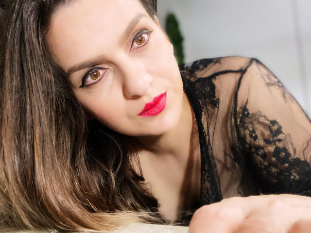 ReveMignoneX has brown eyes, red lipstick, and black lace top, lying with brown hair, looking up with a slight smile. Her tits are partially visible. Her ass is not shown.