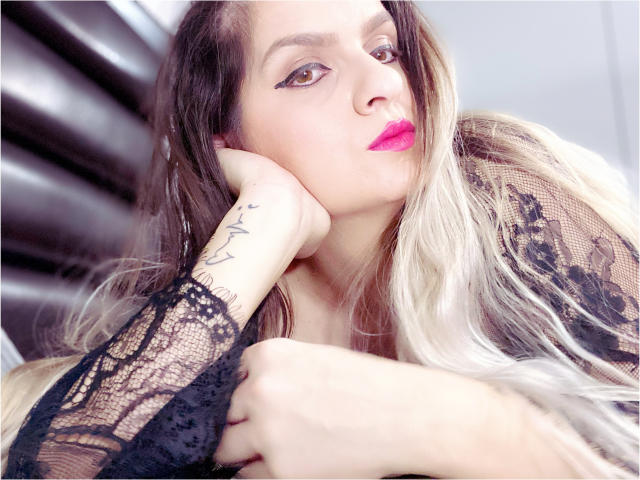 ReveMignoneX with pink lipstick, black lace sleeves, blonde and brown hair, tattoo on right wrist, resting chin on hand, looking up with a slight smile, in a relaxed pose.