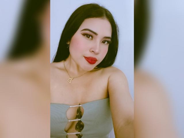 OliviGrace has long black hair, red lipstick, and is wearing a strapless, gray top with a cut-out and a gold heart necklace. Her tits are visible. Mirror reflection.