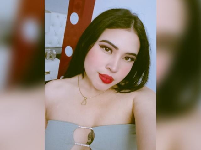 OliviGrace with long black hair, red lipstick, and a strapless grey top with a cutout, wearing a gold necklace, looking at herself in a mirror. Boobs visible.