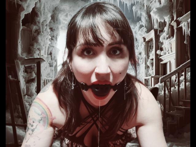 TuttiFrutty, con un mordisque de cuero negro, babeando, tiene ojos grandes y un tatuaje colorido en su brazo derecho. Lleva una blusa de rejilla y está inclinada hacia adelante en una habitación oscura..