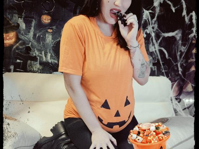 TuttiFrutty in an orange shirt with a pumpkin face, eating candy from a bowl, black leggings, tattoo on right arm, sitting on a white couch, black boots.