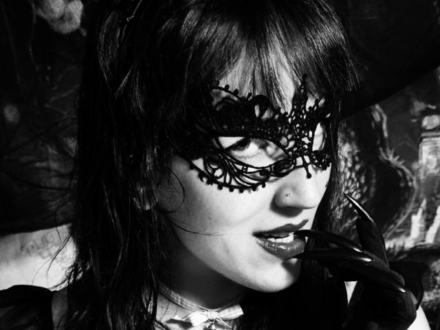 TuttiFrutty's black lace mask covers her eyes, with dark, glossy lipstick and a small nose piercing. She's biting a black glove, her wet, dark hair in her face.