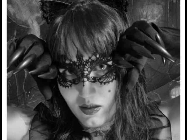 Fotografía en blanco y negro de TuttiFrutty con garras oscuras en la cara, usando una máscara de encaje, lápiz labial oscuro y orejas de gato, con una pequeña peca en la mejilla..