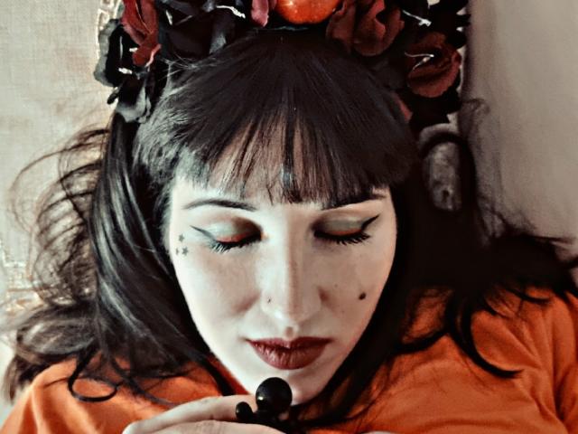 TuttiFrutty with dark hair, wearing an orange shirt, black flower headpiece, and black lipstick, has eyeliner and star tattoos, looking down with eyes closed, holding a black object.