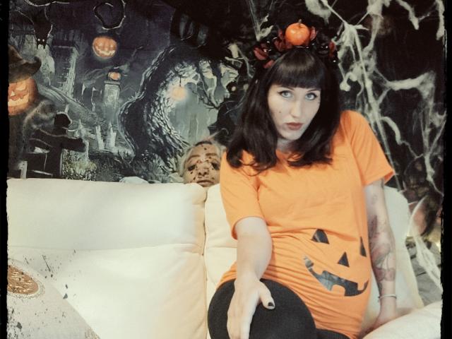 TuttiFrutty with dark hair, orange t-shirt, pumpkin headband, black leggings, tattooed arm, sitting on white couch, pumpkin face on shirt, spooky wallpaper, looking at camera.