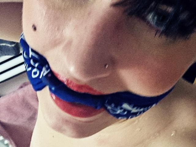 TuttiFrutty's face with a blue bra partially covering her shiny red lips and a small mole on her right cheek. Her skin is pale, and she has dark, short hair.