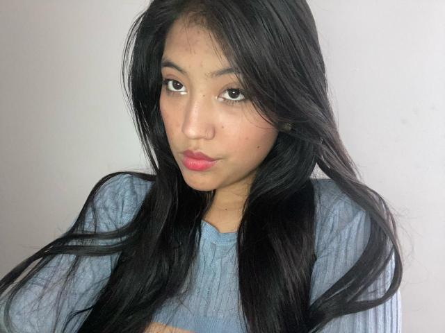 Elizaa has long black hair, brown eyes, and pink lipstick. She's wearing a blue rib-knit shirt, with her tits slightly visible, and a small beauty mark on her cheek.