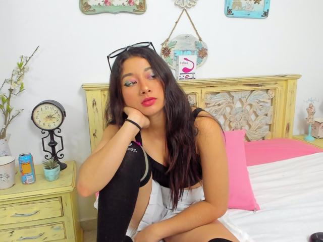 Genesiss with long dark hair, wearing a black and white top, black sock, and glasses on her head, sitting on a bed with pink pillow, looking tired.