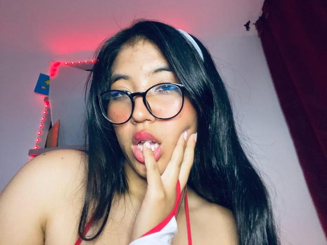 Elizaa, con cabello negro largo, gafas y labios rosados, chupándose el dedo, vistiendo un top blanco con tirantes, una correa roja y una diadema blanca. Una tira de LED roja en el fondo..