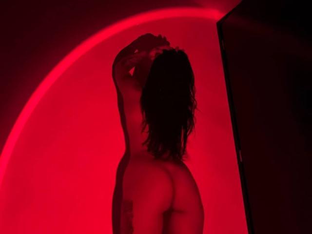 A red-lit photo of Amberv standing naked with arms raised, showcasing her round ass and tattoo on her right side. Her dark hair is messy and she's facing away.