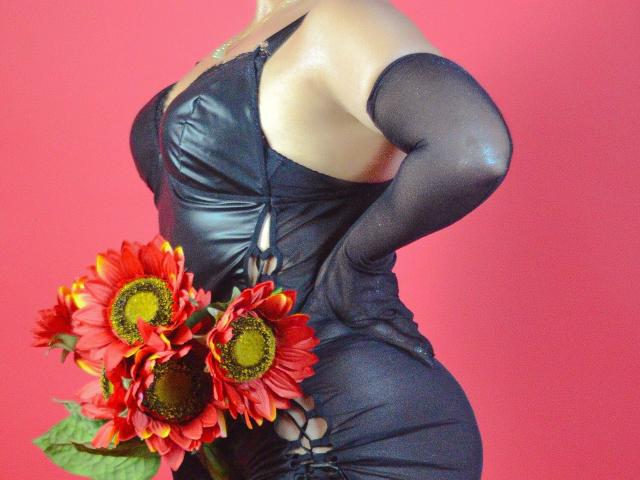 JadeSexiest in a black leather dress with cutouts, holding red sunflowers, showing her curvy ass and big boobs, wearing black mesh sleeves, against a red background.