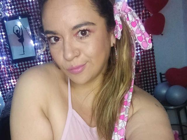 NaugthyCurvy com batom rosa, olhos castanhos, piercing no nariz e um elástico de cabelo rosa com padrão de coração, vestindo um top de alça rosa, mostrando o ombro e a parte superior do peito, sorrindo sutilmente..