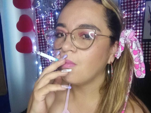 NaugthyCurvy with glasses, nose ring, pink heart bow, and long blonde ponytail, smoking a joint, pink lipstick, white nails, heart-shaped glasses, pink heart-patterned hair tie.