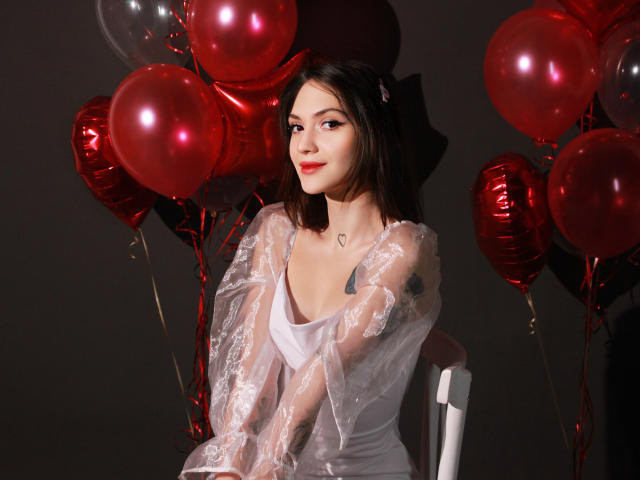 MiraPerez with red balloons, wearing a white lace dress, sitting on a chair, has tattoos on her right arm, dark hair, and a heart necklace, smiling softly.