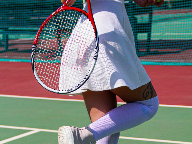 MiraPerez's white tennis skirt flutters, holding a red racket. She's in white socks and sneakers, with a tattoo on her right thigh, on a green and red court.