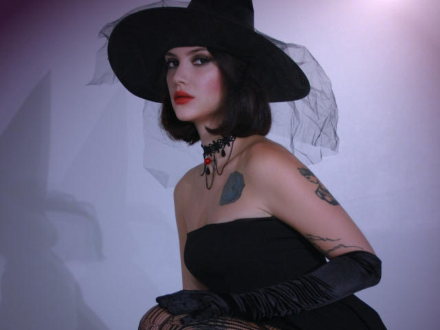 MiraPerez with black witch hat, sheer veil, red lipstick, black strapless dress, black gloves, tattoos, and necklace, looking up with a serious expression.
