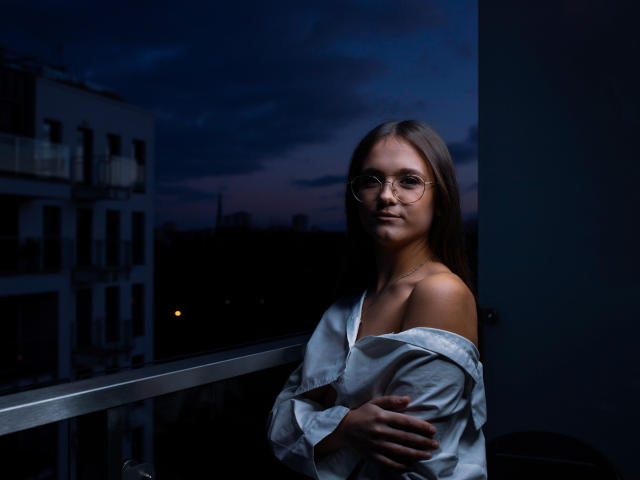 Emily, com óculos, camisa branca com um ombro descoberto, cabelo castanho longo, em pé perto da varanda ao entardecer, segurando a camisa, céu azul escuro, prédios da cidade, sorriso discreto, fofa, sexy..