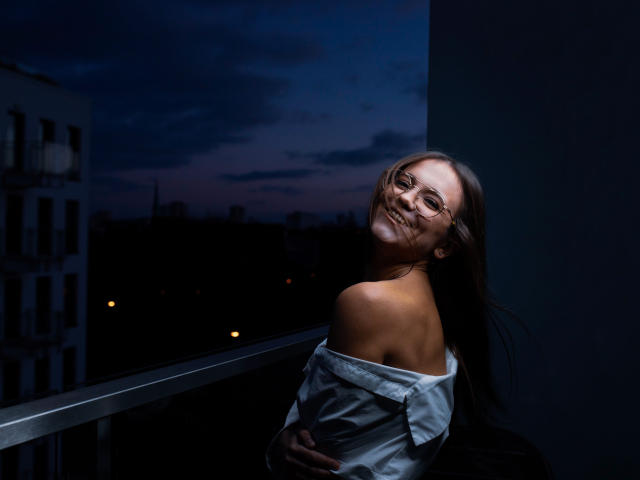 EmilySparkle with glasses, smiling, dark blue evening light, off-shoulder white shirt, long brown hair, bare shoulders, standing on balcony, looking back, hands on shirt, relaxed, cute.