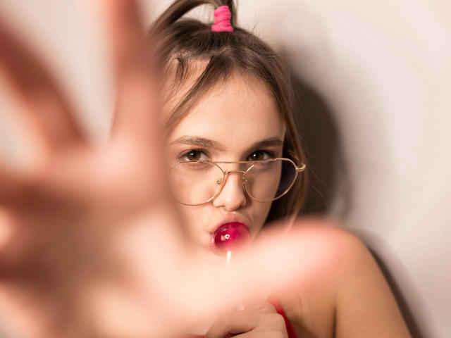 EmilySparkle with pouty pink lips, wearing glasses, has a glossy red lollipop in her mouth, brown hair in a ponytail with a pink scrunchie, and a blurred hand in foreground.