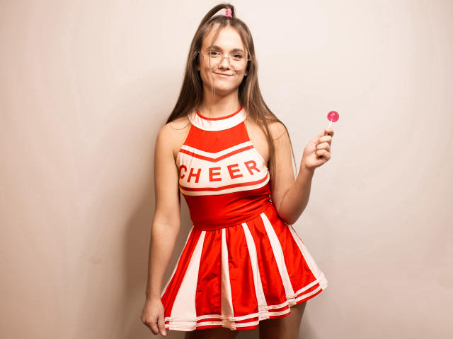 Emily, com cabelos castanhos longos, óculos e um vestido de cheerleader vermelho e branco, segurando um pirulito rosa, sorrindo para a câmera, usando uma presilha rosa no cabelo..