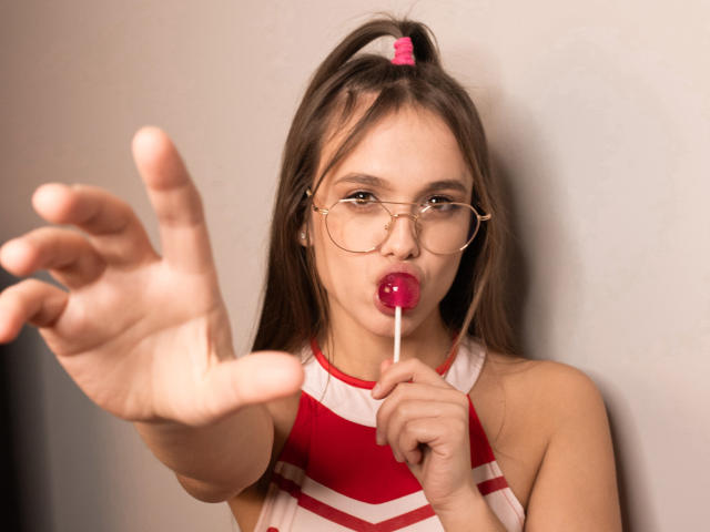 EmilySparkle with long brown hair in a ponytail, gold glasses, and red-and-white top, sucking a pink lollipop, reaching forward with her hand in the foreground.