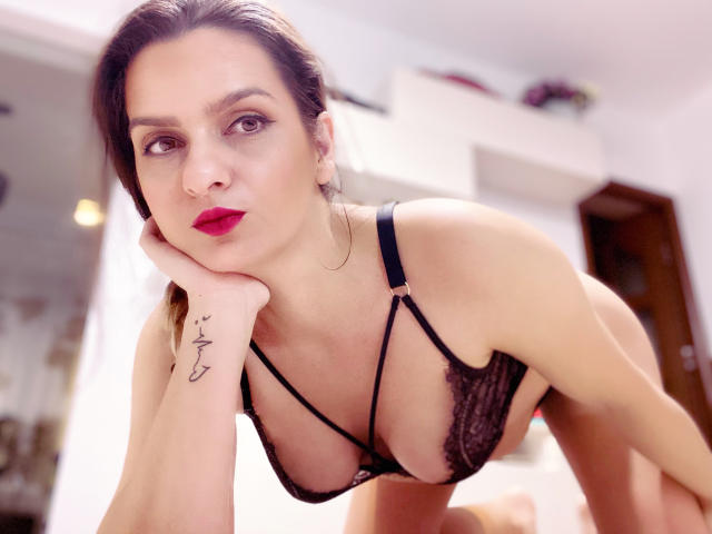 ReveMignoneX with red lipstick, black lace bra, and strappy design, leaning forward, hand on cheek, tattoo on wrist, brown hair, bedroom setting, focused expression, medium shot.