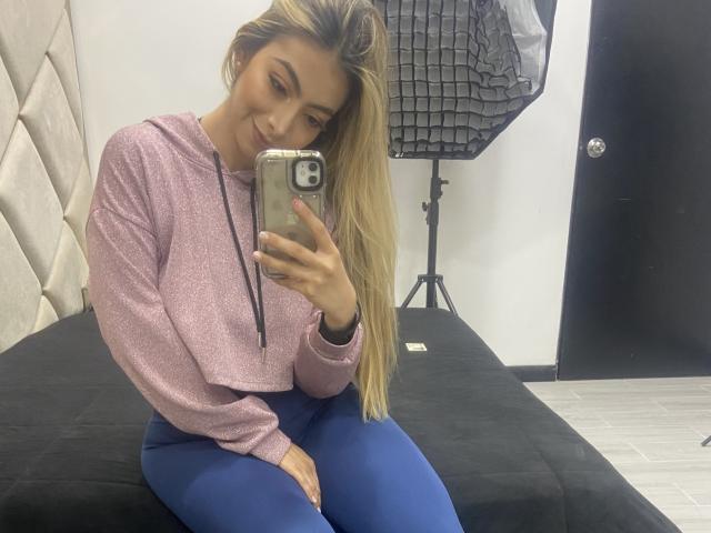 Alessasantini with long blonde hair, pink hoodie, and blue pants, sitting on a black bed, taking a selfie with a smartphone, smiling. Her tits are small and her pussy is hidden.