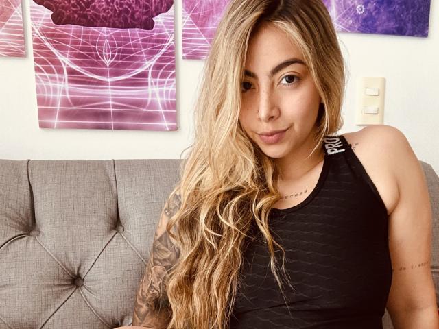 Alessasantini with long blonde hair, black tank top, tattooed arm, and cute face, sitting on a grey couch, giving a slight smirk. Boobs are small and barely visible.