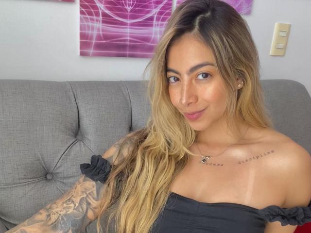 Alessasantini with long blonde hair, black off-shoulder top, tattooed arm, and necklace, sitting on gray couch, smiling, showing tan lines and small tits.