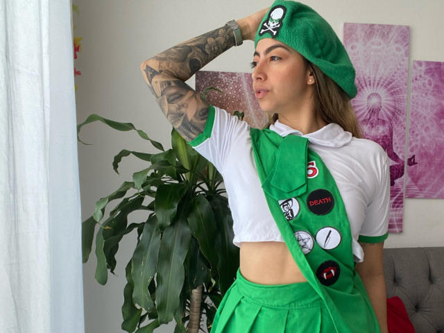 Alessasantini in a green beret, white crop top, and green skirt, with tattoos on her arm, posing with hand on head, wearing a green scarf with buttons.