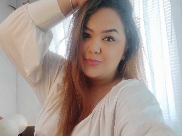 RouseWatsons with long brown hair, pink lipstick, and white shirt, raising her right arm, showing her cute face and small tits. She's smiling slightly, looking at the camera.
