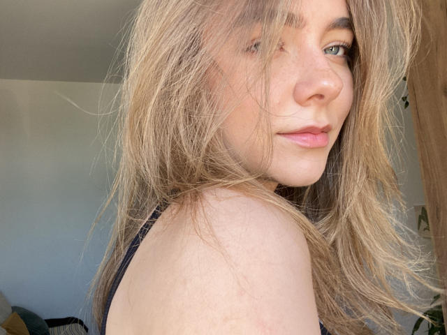 AmyTasty with wavy blonde hair, blue eyes, and pink lips, showing off her bare shoulder and black strap, smiling slightly. Her tits are partially visible, slightly out of frame.