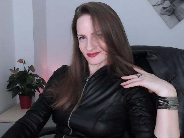 AbyX with long brown hair, red lipstick, black leather top, showing tits, hand on shoulder, black chair, silver bracelet, potted plant on left, smiling, pale skin.