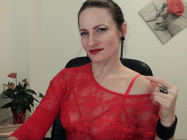AbyX with red lace top, showing bra strap, red lipstick, and black ring, pulling lace down, exposing her tits, in office setting, green plant on desk, smiling.