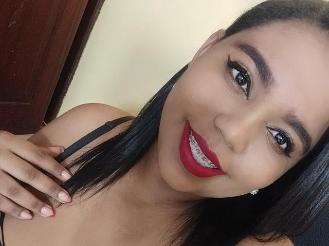 SarahSmity has long black hair, dark eyes, red lipstick, and is smiling with her hand on her shoulder, showing her white braces. She's wearing a black strap top.