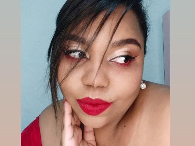SarahSmity has dark hair, red lipstick, and eye makeup with long lashes, wearing a red top, and a diamond earring, touching her chin with a slight smile.