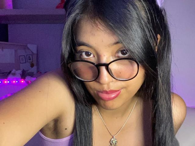Elizaa with black glasses, pink lipstick, and long black hair, wearing a pink top and a necklace with a small pendant, has a cute, pouty face and light brown skin.
