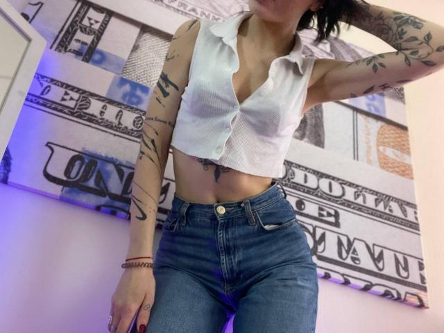 Tsukerberg with tattoos, wearing a white sleeveless button-up shirt exposing her small tits and a flat belly, and high-waisted blue jeans, standing with one arm raised.