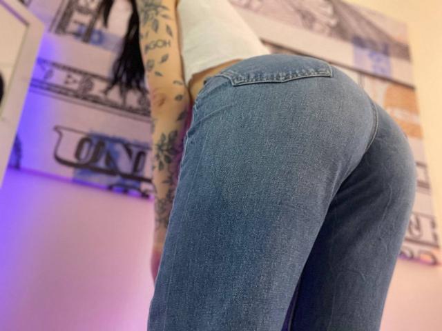 Tsukerberg's tight blue jeans hug her round ass, highlighting her tattooed arm in a white crop top. Her dark hair falls forward, partially visible.
