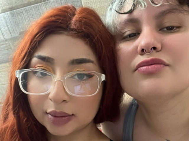 RavenAndEllie with red hair, glasses, and makeup, and Ellie with curly white hair, nose ring, and pink lips, standing close. Ellie's face is on the right.