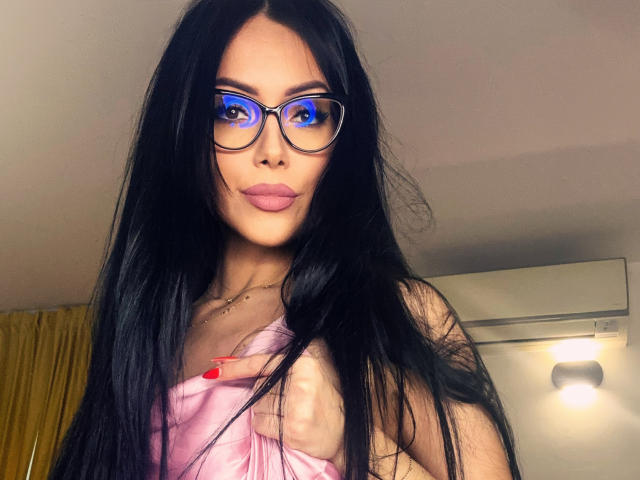 Nadeen with long black hair, blue-lensed glasses, pink pout, and pink dress, showing her tits with red nail, in a softly lit room.