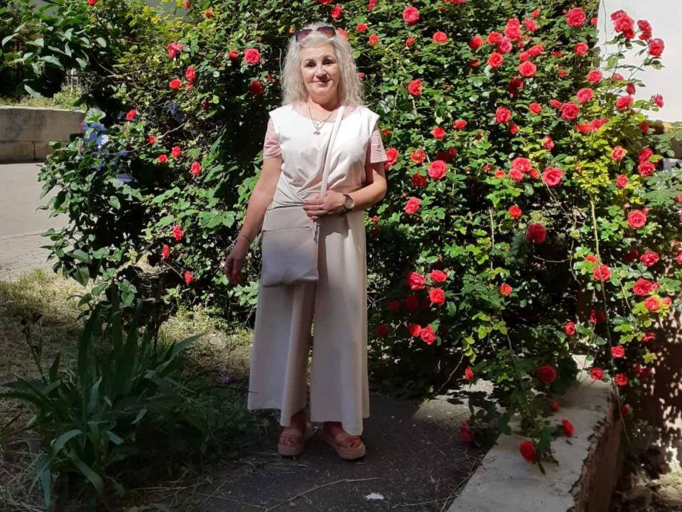Svetulya with blonde hair, white short-sleeve top, beige pants, and pink sandals, standing in front of a rose bush, smiling, holding a gray purse, in a sunny garden.