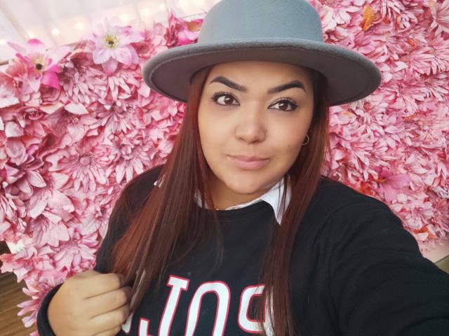 RouseWatson mit roten Haaren, braunen Augen und einem Nasenring, die einen grauen Hut und ein schwarzes Hemd trägt, ihre Haare hochsteckt, lächelt und im Hintergrund rosa Blumen zu sehen sind..