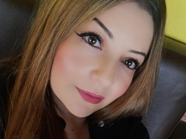 Il viso di NatalyHami, con i suoi lunghi capelli castani, il rossetto rosa e l'eyeliner nero, mette in risalto i suoi grandi occhi marroni e un sorriso adorabile, mentre è seduta su una poltrona in pelle scura..