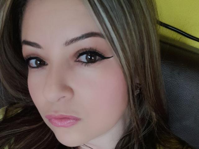 NatalyHami's face with full pink lips, brown eyes, and black eyeliner, has light skin and brown hair with blonde highlights, sitting on a black leather chair.