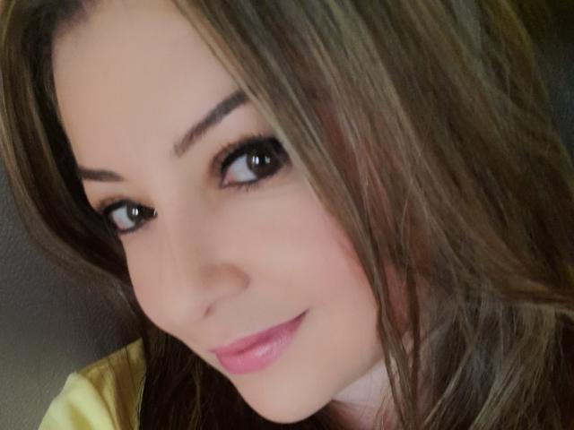 NatalyHami's face, brown eyes, long brown hair, pink lipstick, slight smile, and smooth skin, with a yellow shirt visible. Boobs not shown.