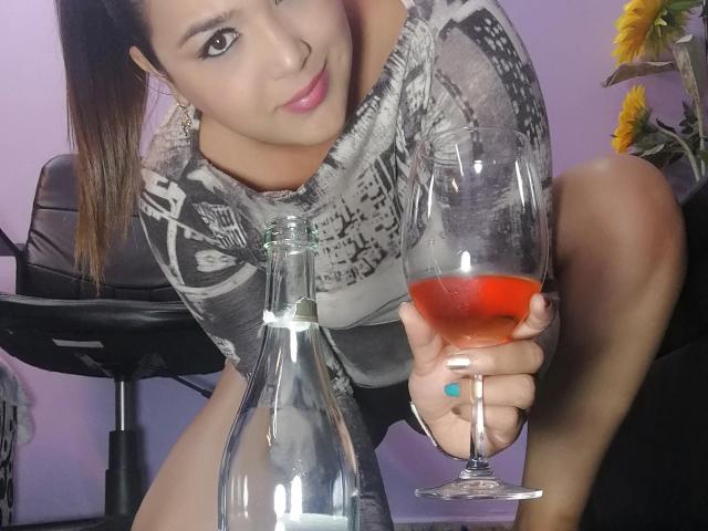 RouseWatsons con capelli scuri, rossetto rosa e unghie bianche, che tiene un cocktail rosso, seduta, indossa una camicia grigia a fantasia e mostra il sedere scoperto, con una bottiglia di vetro..
