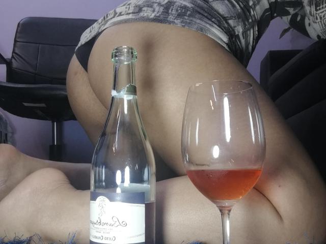 RouseWatsons kniet, wobei ihr Gesäß nach oben zeigt. Sie trägt schwarze Unterwäsche und ein graues Oberteil mit Muster. Im Vordergrund befinden sich eine Flasche Weißwein und ein Glas Roséwein..